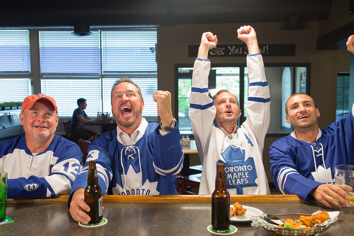 4 adult men wearing Toronto Maple Leaf jersey, drinking beer and cheering for their team.