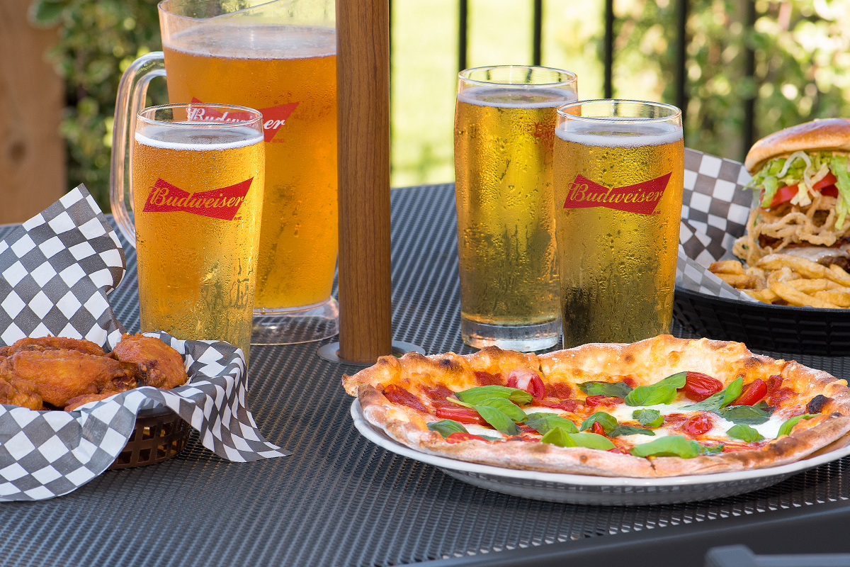 Outdoor patio table with a pitcher of cold beer, pizza, chicken wings and a burger and fries